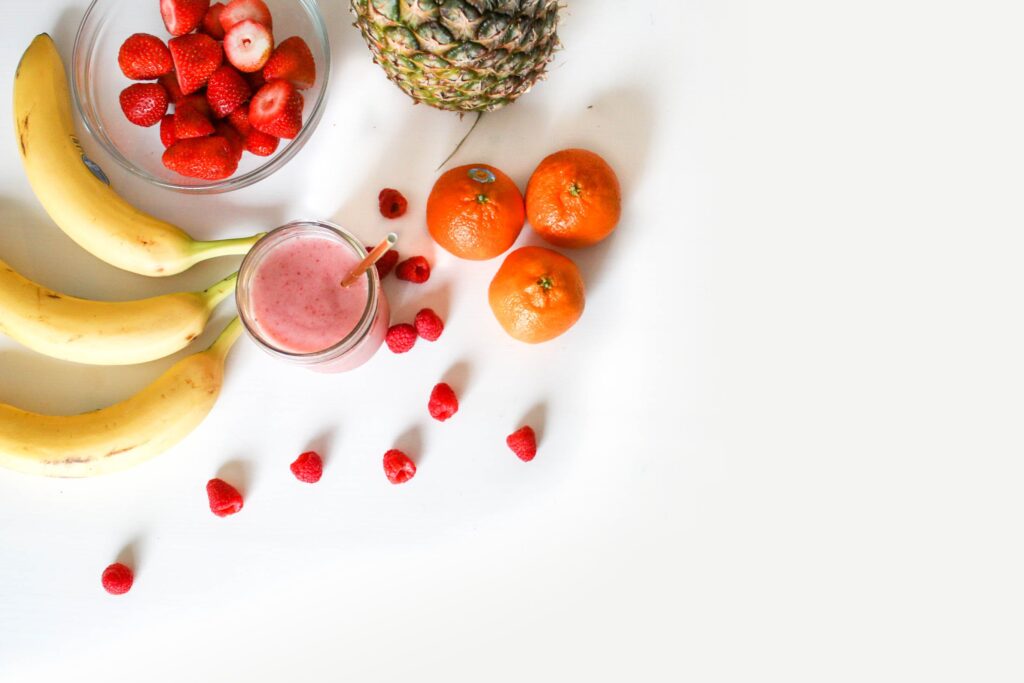 A variety of fresh fruits, including strawberries in a bowl, bananas, oranges, raspberries, and a pineapple, arranged neatly next to a glass of pink smoothie on a white background.